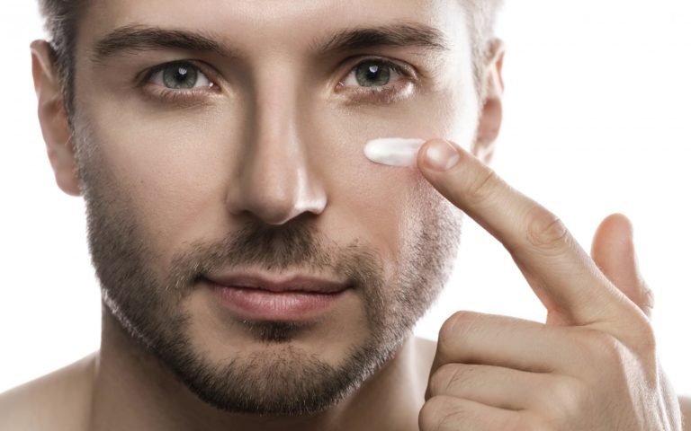 Ein Mann trägt eine Creme auf seine Wange auf, während er in die Kamera schaut. Hintergrund ist neutral.