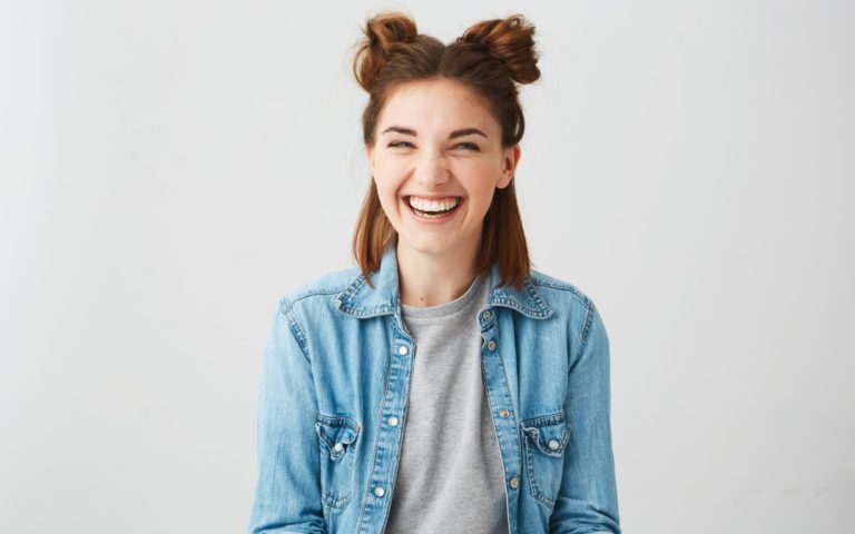 Lachende junge Frau mit Duttfrisur, trägt eine Jeansjacke über einem grauen T-Shirt, vor einem hellen Hintergrund.