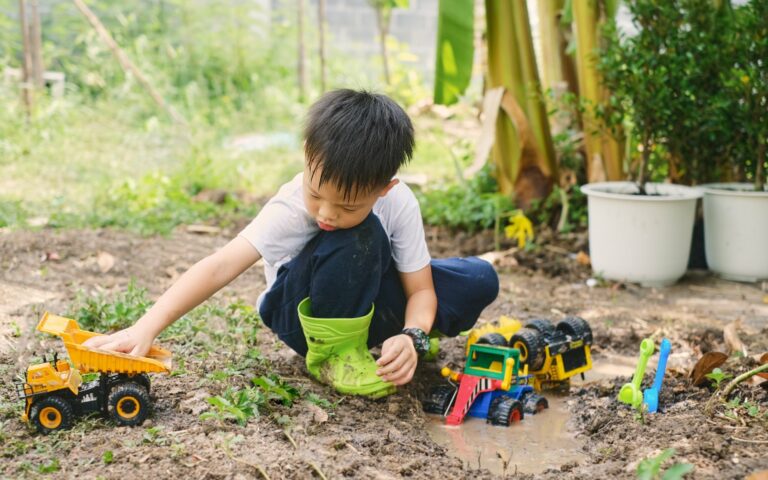 Ein Junge im Garten spielt mit verschiedenen Spielzeugfahrzeugen, darunter ein Lkw und ein Bagger, während er in matschigem Boden sitzt.
