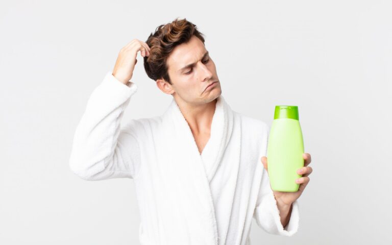 Ein Mann in einem weißen Bademantel hält eine grüne Shampoo-Flasche und sieht nachdenklich aus.