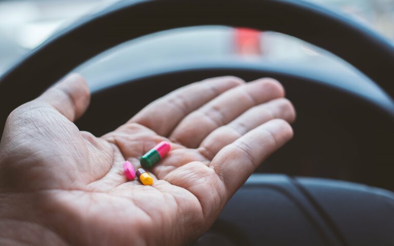 Eine Hand hält bunte Kapseln über dem Lenkrad eines Fahrzeugs.