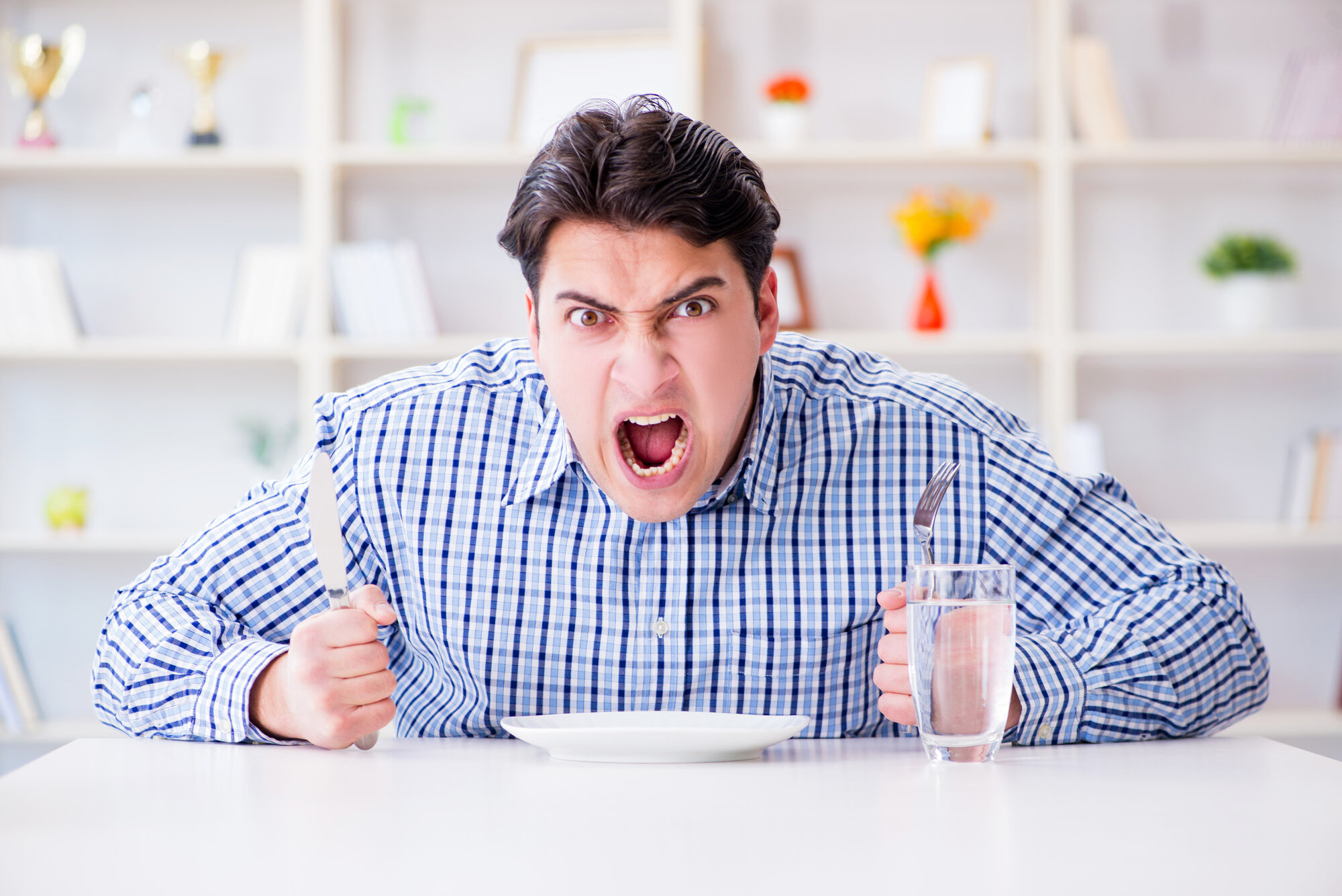 Ein wütender Mann hält ein Messer und eine Gabel über einem leeren Teller, während er an einem Tisch sitzt.