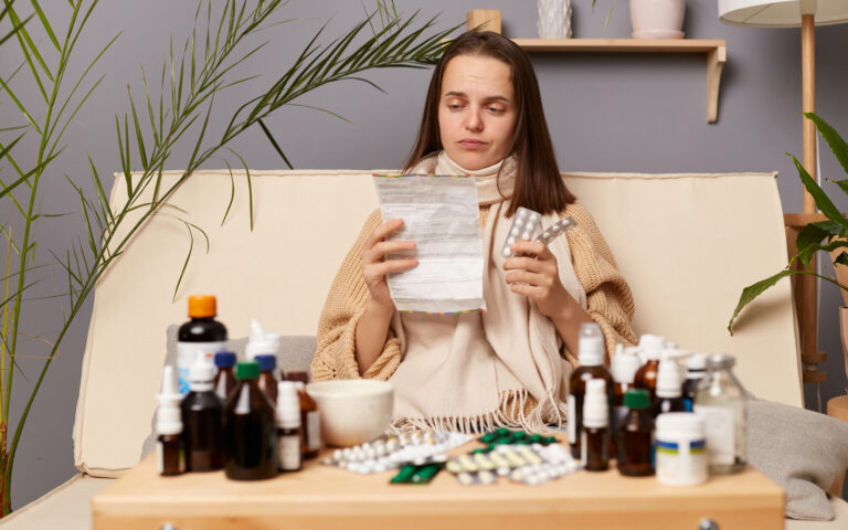 Eine kranke Frau sitzt auf einem Sofa, umgeben von verschiedenen Medikamenten und hält einen Zettel sowie eine Medikamentenpackung in der Hand.