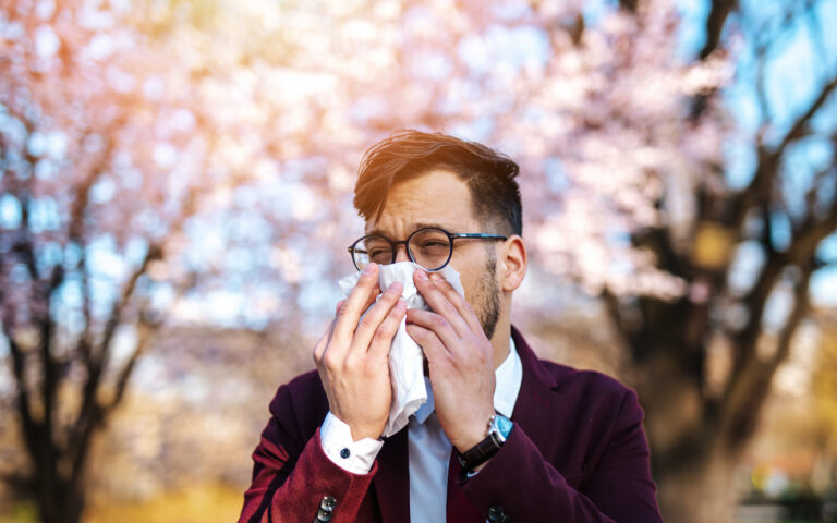 Ein Mann in einem Anzug benutzt ein Taschentuch, um sich die Nase zu putzen, vor blühenden Bäumen im Hintergrund.