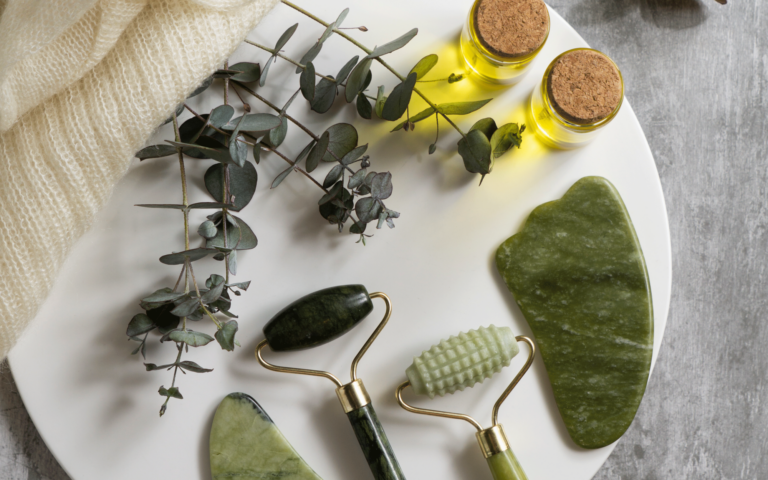 Ein weißer Teller mit drei Jade-Rollern, einem gua sha Stein, zwei Fläschchen ätherischem Öl und Eukalyptuszweigen.