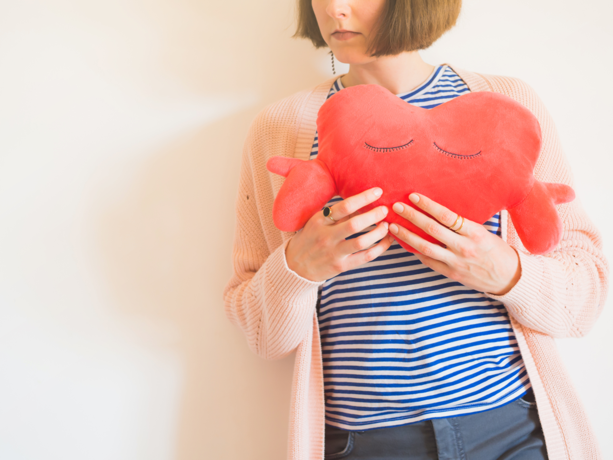Herzinfarkt_Frauen.png Eine Person hält ein rotes herzförmiges Kissen mit geschlossenen Augen, gekleidet in gestreiftes Oberteil und eine helle Strickjacke.