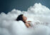 Frau in einem weißen Kleid, die entspannt auf weichen, grauen Wolken liegt, mit geschlossenen Augen und einem friedlichen Ausdruck.