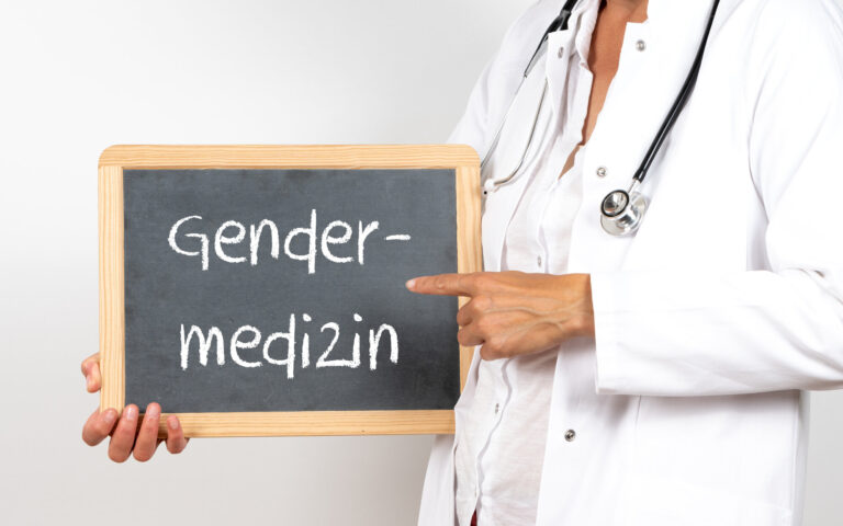 Eine Person in Arztkleidung hält eine Tafel mit der Aufschrift "Gendermedizin".