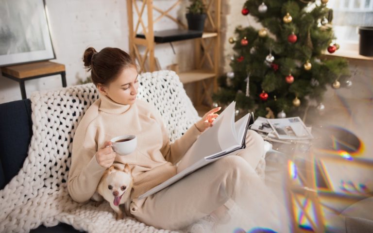 Eine Frau in bequemer Kleidung sitzt auf einem Sofa, liest in einem Buch und hält eine Tasse Kaffee, während ein Hund bei ihr sitzt.