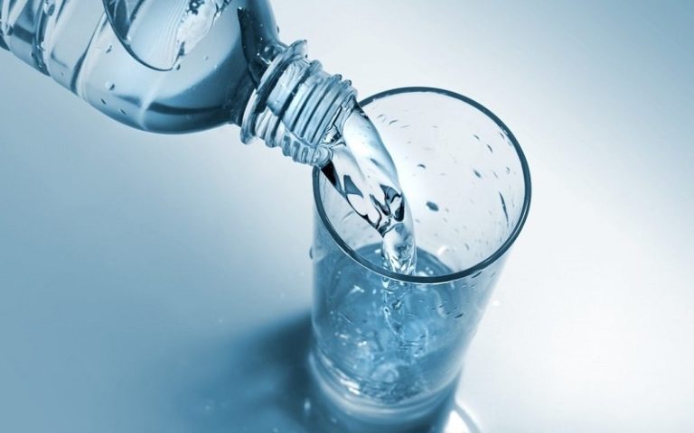 Eine Wasserflasche schüttet frisches Wasser in ein Glas, das auf einer hellen, blauen Oberfläche steht.