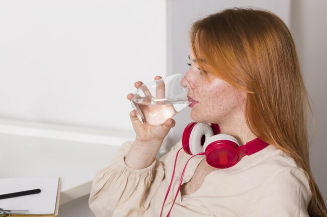 Eine Person mit roten Kopfhörern trinkt aus einem Glas Wasser und sitzt an einem Schreibtisch.