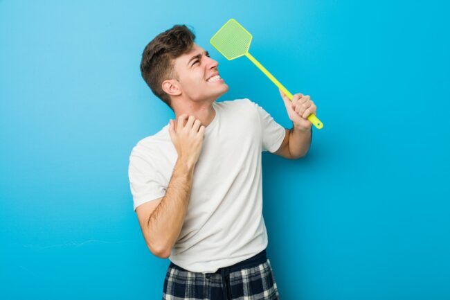 Junger Mann in einem weißen T-Shirt hält ein grünes Insektenspray mit einem lächelnden Gesicht vor einer blauen Wand.