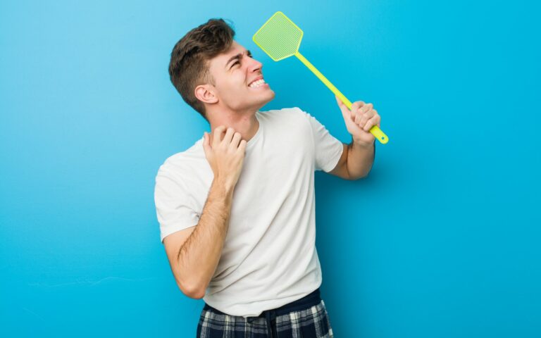 Junger Mann in einem weißen T-Shirt hält ein grünes Insektenspray mit einem lächelnden Gesicht vor einer blauen Wand.