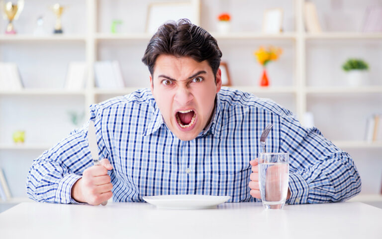 Ein wütender Mann hält ein Messer und eine Gabel über einem leeren Teller, während er an einem Tisch sitzt.