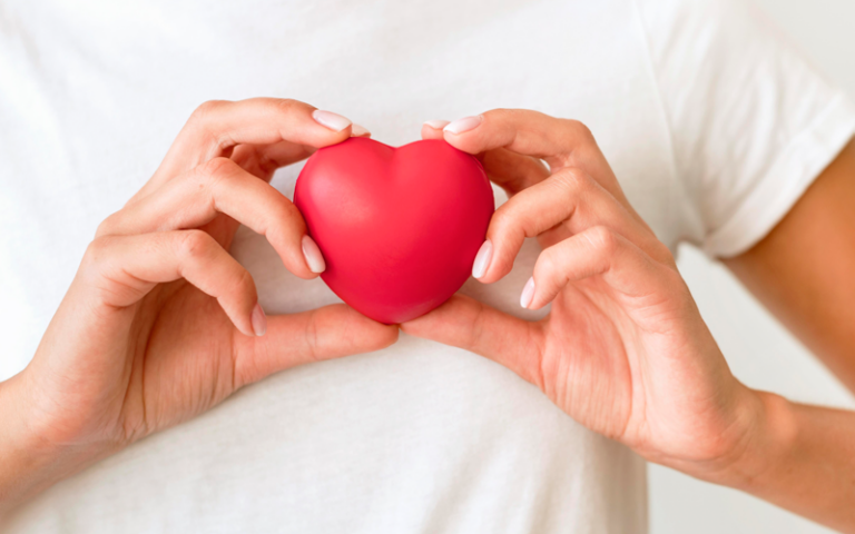 Eine Person hält ein rotes Herzen in beiden Händen vor einem hellen Hintergrund.