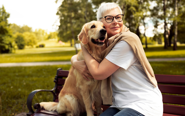 Eine Frau mit kurzen, blonden Haaren und Brille umarmt einen golden retriever auf einer Bank im Park.