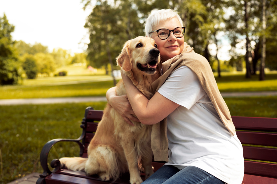 Hunde-als-Seelentroester.png Eine Frau mit kurzen, blonden Haaren und Brille umarmt einen golden retriever auf einer Bank im Park.