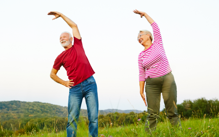 Ein älteres Paar führt Dehnübungen im Freien auf einer Wiese durch, umgeben von Landschaft und Hügeln.