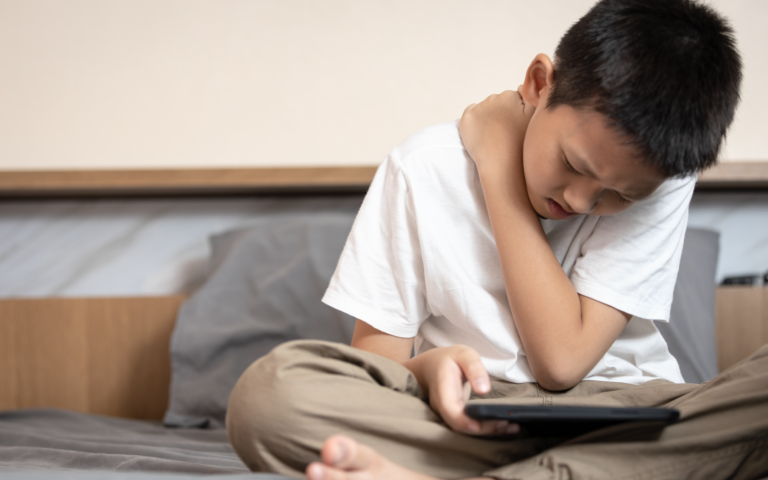 Ein Junge sitzt auf einem Bett, hält ein Tablet und fasst sich schmerzverzerrt an den Hals.
