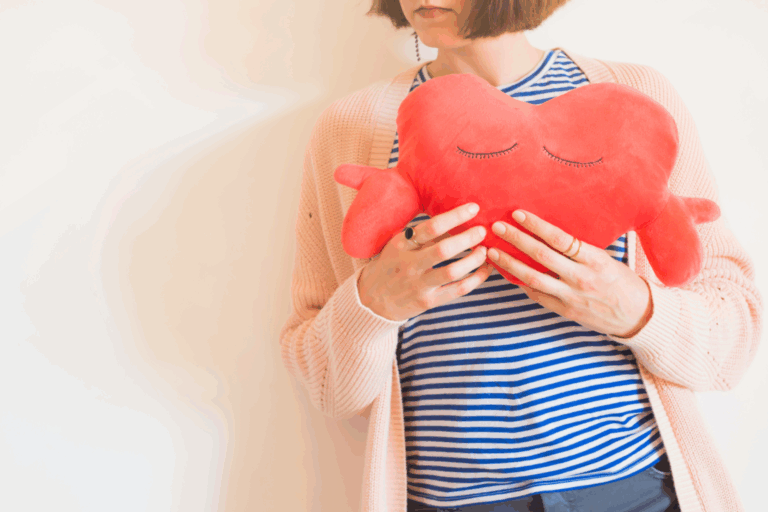 Eine Person hält ein rotes herzförmiges Kissen mit geschlossenen Augen, gekleidet in gestreiftes Oberteil und eine helle Strickjacke.