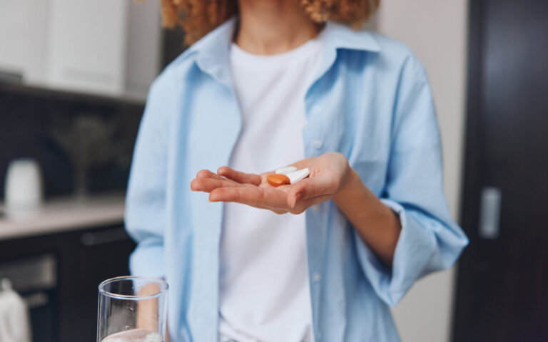 Eine Person hält zwei Kapseln in einer Hand, während in der anderen ein Glas Wasser steht, in einer modernen Küche.