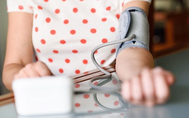 Eine Person mit einem Blutdruckmessgerät am Arm, das auf einem Tisch liegt. Im Hintergrund ein unscharfer Raum.
