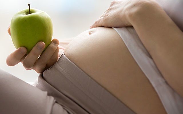 Eine schwangere Person hält einen grünen Apfel und berührt sanft ihren Bauch.