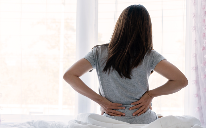 Eine Person mit langen Haaren sitzt auf einem Bett und hält sich den Rücken vor einem Fenster.