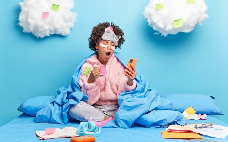 Eine Person in einem blauen Bett, umhüllt von einer Decke, schaut müde auf ihr Handy und hält eine Tasse in der Hand. Wolken mit Haftnotizen im Hintergrund.