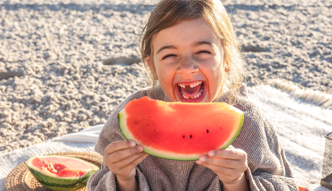 Ein lachendes Kind hält ein großes Stück Wassermelone in der Hand, umgeben von einer sommerlichen Picknick-Atmosphäre.
