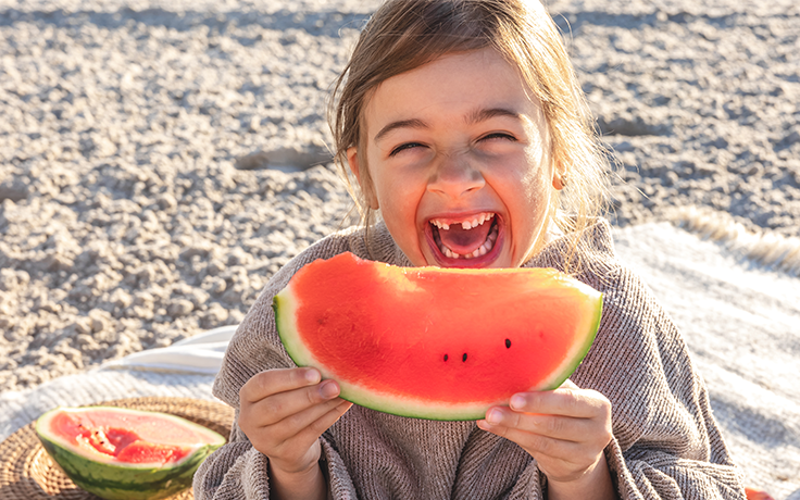 Ein lachendes Kind hält ein großes Stück Wassermelone in der Hand, umgeben von einer sommerlichen Picknick-Atmosphäre.