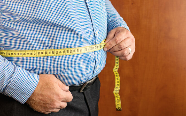 Mann misst seinen Bauchumfang mit einem gelben Maßband, trägt ein kariertes Hemd und dunkle Hose.