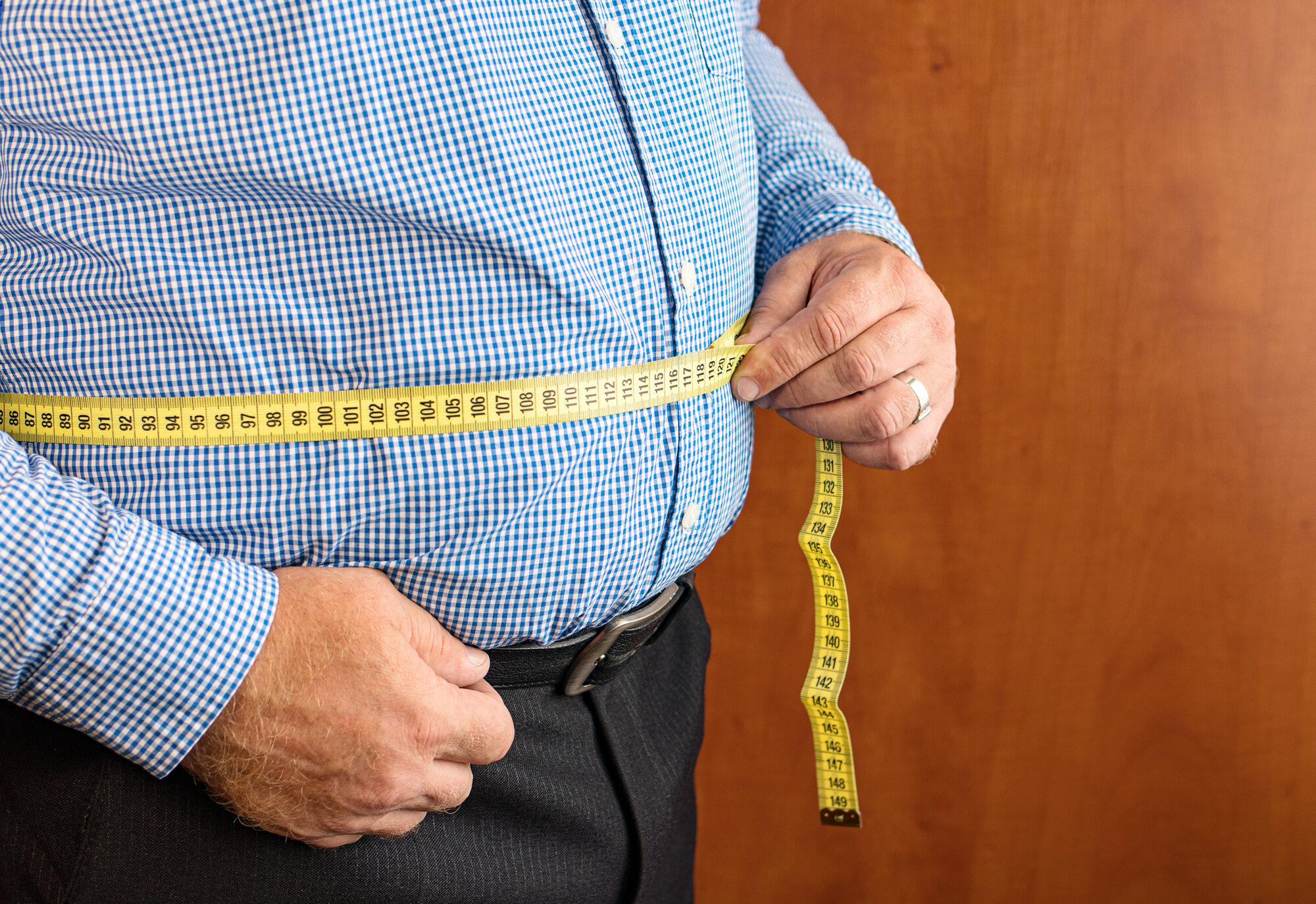 adipositas.jpg Mann misst seinen Bauchumfang mit einem gelben Maßband, trägt ein kariertes Hemd und dunkle Hose.