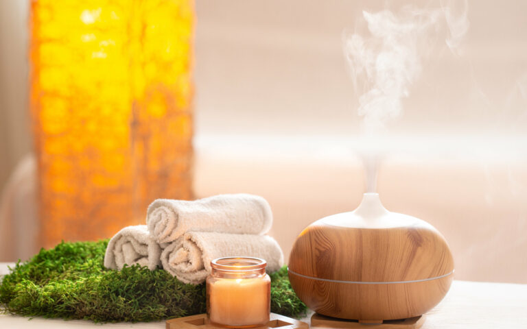 Ein Aromadiffusor aus Holz mit aufsteigendem Dampf, umgeben von Handtüchern, einer Kerze und grünem Moos.