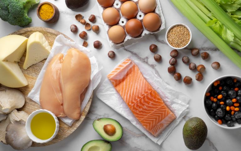 Verschiedene Lebensmittel wie Hähnchenbrust, Lachs, Käse, Eier, Avocado und Gemüse auf einem Tisch.