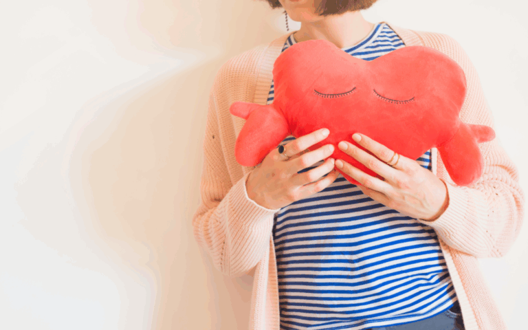 Eine Person hält ein rotes herzförmiges Kissen mit geschlossenen Augen, gekleidet in gestreiftes Oberteil und eine helle Strickjacke.